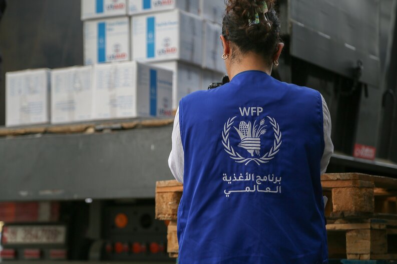 Eine Frau, die eine WFP-Weste trägt, von hinten gesehen, steht vor Lebensmittelpaketen. © WFP/ Mohammad Awadh