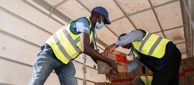 WFP-Mitarbeiter in Warnwesten laden Kisten mit humanitären Hilfsgütern von einem Lkw ab und transportieren die Kartons im Rahmen der Logistikmaßnahmen von Hand weiter.