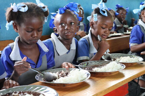 Foto: WFP/Alexis Masciarelli. Schülerinnen und Schüler im Alter von 5 bis 7 Jahren essen eine warme Mahlzeit in ihrem Klassenzimmer der Grundschule Baptiste Salem in Dondon, an der 600 Kinder registriert sind. Die Mahlzeiten werden täglich mit Reis und Bohnen zubereitet – Lebensmittel, die über das WFP-Programm für lokal produzierte Schulmahlzeiten von lokalen Produzenten gekauft werden.