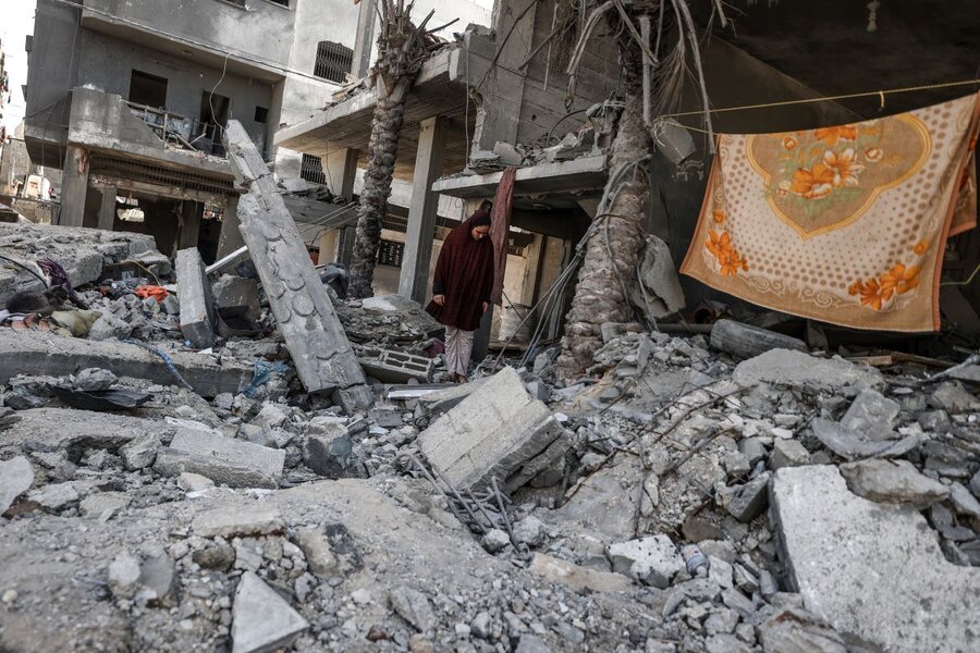 a woman in a dark coat and head scarf walks among rubble in Gaza