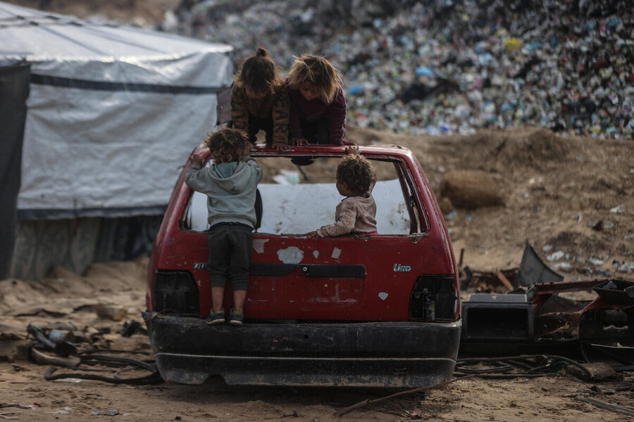 Four children play in the window-less shell of an abandoned car near what looks like a landfill site