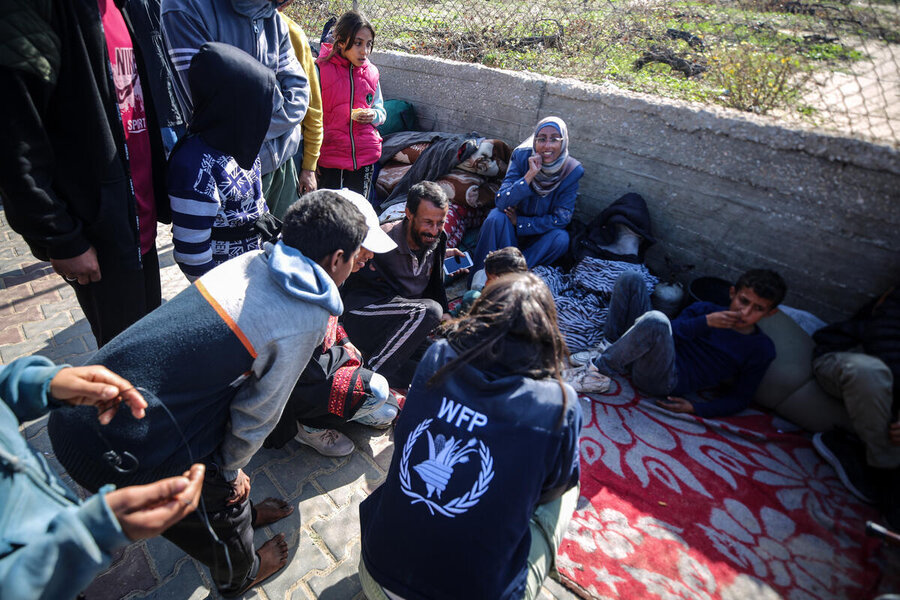 A group of people, including children, sit on blankets against a concrete wall. A WFP worker in a navy jacket with the agency’s logo kneels, engaging with them